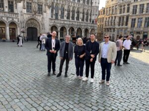 Na zdjęciu widocznych jest pięć osób stojących na wybrukowanym placu w zabytkowym centrum Brukseli. To pracownicy Katedry oraz przedstawiciele Santander Bank Polska, uchwyceni podczas zwiedzania. W tle znajduje się imponujący, historyczny budynek o bogato zdobionej fasadzie, z łukowatymi oknami i rzeźbami. Wokół poruszają się inni przechodnie, a słoneczna pogoda dodaje scenie turystycznego i przyjaznego charakteru.