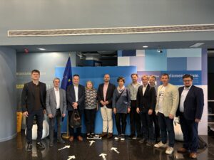 Na zdjęciu znajduje się grupa jedenastu osób stojących wewnątrz budynku, na tle ścianki z flagą Unii Europejskiej. To pracownicy Katedry oraz przedstawiciele Santander Bank Polska, pozujący do zdjęcia w Parlamentarium – centrum dla odwiedzających Parlament Europejski. Wszyscy są elegancko ubrani, część osób trzyma ręce w kieszeniach lub splecione przed sobą. W tle widoczne są napisy oraz elementy dekoracyjne związane z instytucjami europejskimi. Cała scena ma miejsce w nowoczesnym wnętrzu z marmurową podłogą.