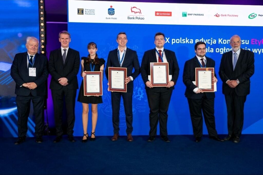 Zdjęcie grupowe laureatów i organizatorów X polskiej edycji Konkursu 'Etyka w finansach'. Pięć osób w centralnej części kadru trzyma oprawione dyplomy, stojąc na tle niebieskiej ścianki z logotypami partnerów konkursu, m.in. Związku Banków Polskich, Banku Pekao, PKO Banku Polskiego, Santander Bank Polska, BNP Paribas i innych. Laureaci ubrani są formalnie – kobieta w eleganckiej czarnej sukience, mężczyźni w garniturach. Po bokach stoją czterej starsi mężczyźni – członkowie jury oraz przedstawiciele instytucji wspierających.