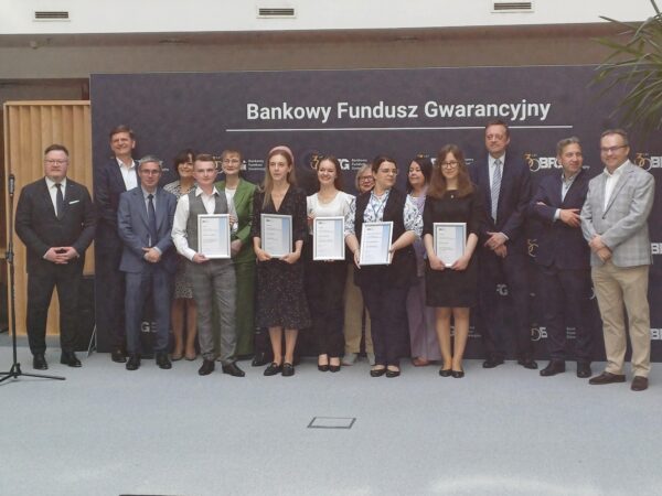 Grupowe zdjęcie uczestników i organizatorów wydarzenia z okazji wręczenia nagród lub certyfikatów przez Bankowy Fundusz Gwarancyjny. W pierwszym rzędzie stoi sześć osób trzymających dyplomy, za nimi grupa przedstawicieli w formalnych strojach. W tle ścianka z logo Bankowego Funduszu Gwarancyjnego.