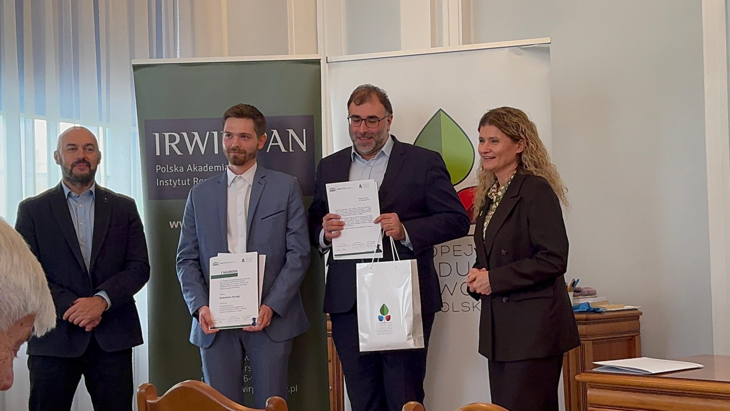 Zdjęcie przedstawia formalne wydarzenie odbywające się w pomieszczeniu, gdyż chodzi o ceremonię wręczenia nagród lub wyróżnień. W pierwszym planie stoją cztery osoby w eleganckich strojach — jedna z nich trzyma certyfikat i torbę z logo, a pozostali patrzą w jej stronę z uśmiechem. W tle widoczne są dwa banery z logo i nazwami „IRW” oraz „Polska Akademia Nauk”, co wskazuje na charakter naukowy lub instytucjonalny wydarzenia. Atmosfera jest uroczysta i profesjonalna, podkreślająca moment uznania lub osiągnięcia.