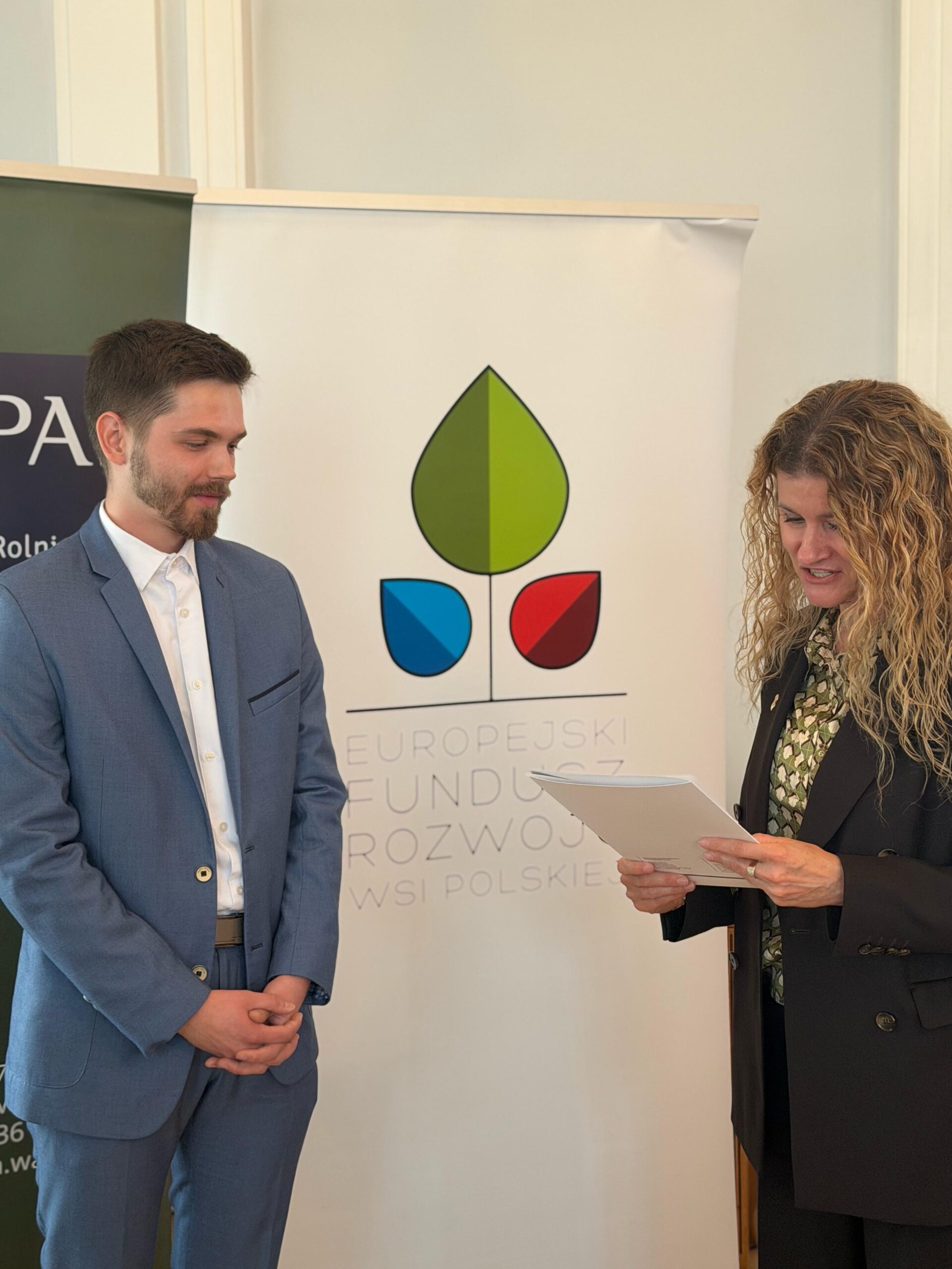 Na zdjęciu widać mężczyznę i kobietę stojących w pomieszczeniu podczas formalnego wydarzenia lub konferencji. Mężczyzna po lewej ma jasnoniebieski garnitur i białą koszulę, trzyma dłonie przed sobą i z lekkim uśmiechem patrzy na kobietę. Kobieta po prawej ma długie, kręcone blond włosy, ciemną marynarkę i trzyma w rękach plik dokumentów, jakby prowadziła prezentację lub rozmowę. W tle widoczny jest baner z logo i napisem „Europejski Fundusz Rozwoju Wsi Polskiej”, co sugeruje, że wydarzenie dotyczy rozwoju obszarów wiejskich. Atmosfera jest profesjonalna i oficjalna, oświetlenie jasne i równomierne.