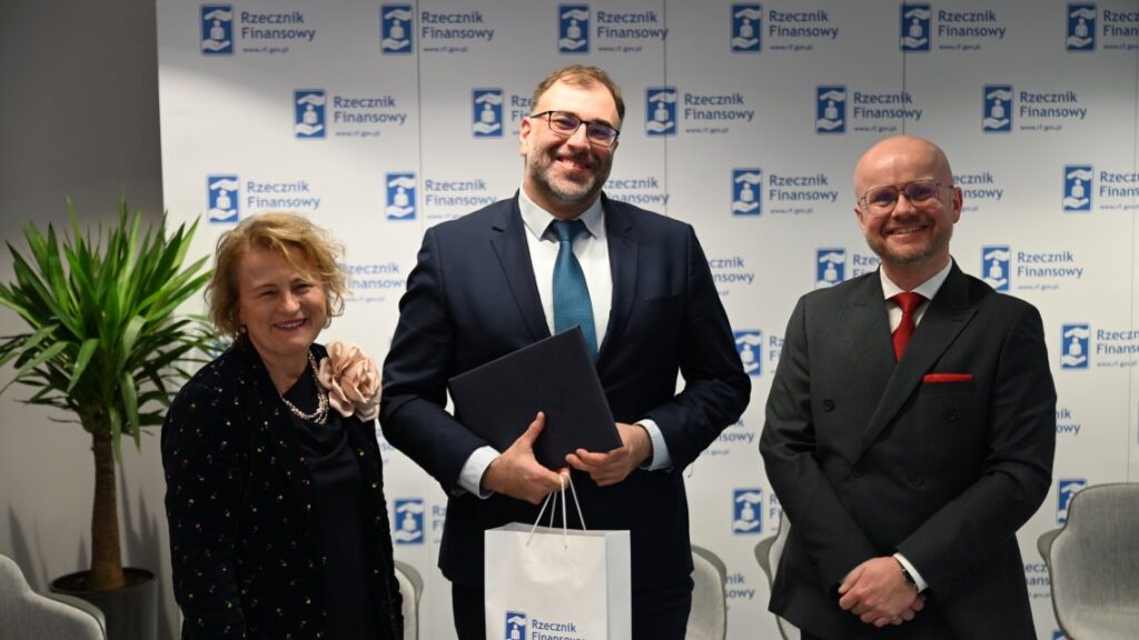 Zdjęcie przedstawia trzy osoby stojące obok siebie podczas formalnego wydarzenia, prawdopodobnie konferencji prasowej lub oficjalnego ogłoszenia, zorganizowanego przez instytucję Rzecznik Finansowy. Postacie znajdują się na pierwszym planie i pozują do zdjęcia na tle białego baneru z powtarzającym się logo oraz nazwą organizacji, co podkreśla profesjonalny i oficjalny charakter sytuacji. Po lewej stronie stoi kobieta z krótkimi, kręconymi włosami, ubrana w ciemny strój z wzorzystą bluzką i naszyjnikiem, uśmiechnięta i zwrócona w stronę aparatu. W centrum znajduje się mężczyzna w ciemnym garniturze z niebieskim krawatem, trzymający w rękach teczkę lub dokument, co sugeruje prezentację lub przekazanie oficjalnego materiału, a jego postawa i uśmiech wskazują, że jest w centrum uwagi. Po prawej stronie stoi drugi mężczyzna w ciemnym garniturze z czerwonym krawatem, z rękami splecionymi przed sobą, również uśmiechnięty i spokojny. Cała scena rozgrywa się we wnętrzu, najprawdopodobniej w sali konferencyjnej lub biurze przystosowanym do oficjalnych wystąpień, z jasnym, równomiernym oświetleniem. Atmosfera zdjęcia jest pozytywna, profesjonalna i uporządkowana, a wyraz twarzy uczestników sugeruje satysfakcję oraz rangę wydarzenia.