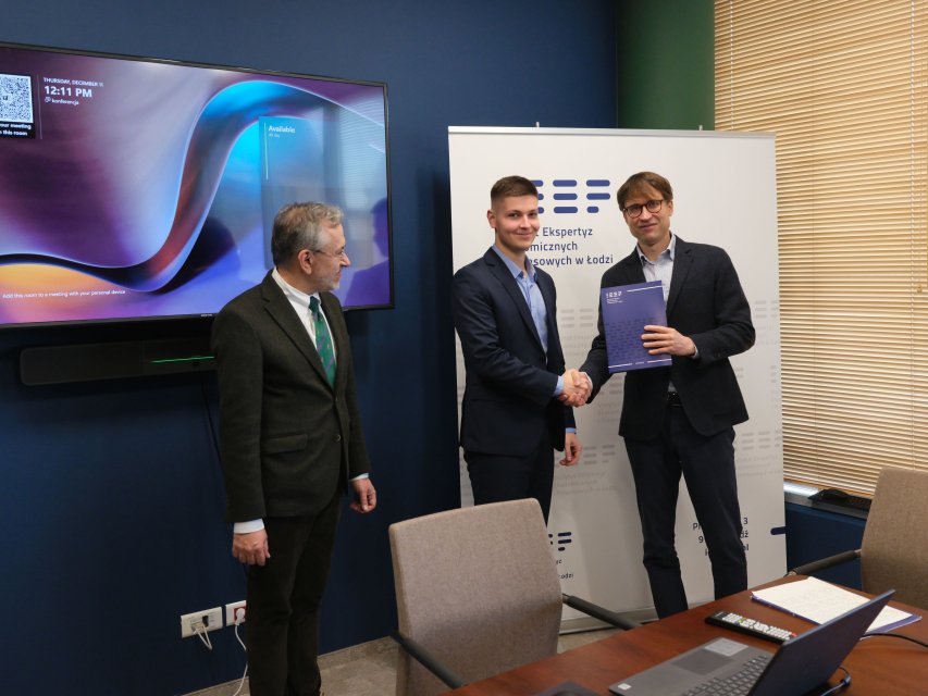 Zdjęcie przedstawia profesjonalne wnętrze, prawdopodobnie biuro lub salę konferencyjną, w którym trzech mężczyzn uczestniczy w formalnym spotkaniu. Na pierwszym planie widoczne jest biurko z otwartym laptopem, kilkoma dokumentami oraz niewielkim urządzeniem przypominającym pilot, co podkreśla roboczy charakter miejsca. Dwaj mężczyźni znajdujący się w centrum kadru podają sobie ręce, co sugeruje zawarcie porozumienia, zakończenie rozmów lub potwierdzenie współpracy; obaj są ubrani w stroje biznesowe, a jeden z nich trzyma w drugiej ręce teczkę lub broszurę z dokumentami. Trzeci mężczyzna, stojący po lewej stronie, obserwuje ten gest, sprawiając wrażenie świadka lub uczestnika spotkania. W tle znajduje się duży ekran wyświetlający kolorowy, abstrakcyjny obraz przypominający slajd prezentacji lub interfejs cyfrowy, na którym widoczny jest także znacznik czasu „12:11 PM”. Obok ekranu częściowo widoczny jest baner z polskim tekstem zawierającym słowa odnoszące się do ekspertów i zagadnień ekonomicznych, co sugeruje kontekst zawodowy lub ekspercki wydarzenia. Cała scena ma formalny i profesjonalny charakter, a atmosfera jest spokojna, rzeczowa i skupiona na sprawach biznesowych lub organizacyjnych.