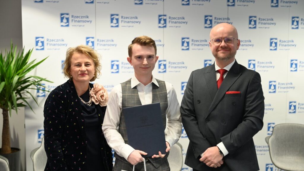 Zdjęcie przedstawia trzy osoby stojące obok siebie podczas formalnego wydarzenia, prawdopodobnie ceremonii wręczenia nagród lub spotkania zawodowego. Po lewej stronie znajduje się kobieta o krótkich, kręconych włosach, ubrana w czarną sukienkę ozdobioną kolorowymi groszkami oraz kwiatowym dodatkiem na ramieniu. W centrum stoi młody mężczyzna w białej koszuli i szarej kamizelce, trzymający w dłoniach certyfikat lub dokument, co sugeruje moment wyróżnienia lub nagrodzenia. Po prawej stronie widoczny jest mężczyzna w ciemnym garniturze z czerwoną krawatą, uśmiechający się i patrzący wprost w obiektyw aparatu. W tle znajduje się duży baner z powtarzającym się napisem i logotypem „Rzecznik Finansowy”, co wskazuje na oficjalny charakter wydarzenia związanego z instytucją finansową. Scena rozgrywa się we wnętrzu, prawdopodobnie w sali konferencyjnej lub holu, z widocznymi elementami wyposażenia, takimi jak krzesła oraz doniczka z rośliną po lewej stronie, która przełamuje formalny charakter otoczenia. Atmosfera zdjęcia jest jednocześnie oficjalna i celebracyjna, a uśmiechy uczestników sugerują pozytywny, dumny moment związany z osiągnięciem lub wyróżnieniem.