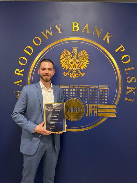Na zdjęciu widoczny jest mężczyzna pozujący z oprawionym certyfikatem, ubrany w jasnoniebieski garnitur i białą koszulę. W dłoniach trzyma dyplom z napisem „DYPLOM”. Stoi przed ciemnoniebieską ścianą z dużym złotym emblematem Narodowego Banku Polskiego. Logo zawiera okrągły napis „Narodowy Bank Polski”, wizerunek orła oraz stylizowany obraz budynku, a także litery „NBP” umieszczone w okręgu. Mężczyzna uśmiecha się, co sugeruje pozytywny i uroczysty charakter sytuacji. Cała scena ma profesjonalny charakter i może być związana z wyróżnieniem lub osiągnięciem w Narodowym Banku Polskim.