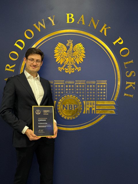Na zdjęciu widoczny jest mężczyzna w okularach, ubrany w białą koszulę, czarną marynarkę i czarne spodnie, który trzyma oprawiony dyplom. Stoi przed niebieską ścianą z dużym, złotym, okrągłym emblematem Narodowego Banku Polskiego. Logo zawiera napis „Narodowy Bank Polski” umieszczony w górnej części okręgu, wizerunek orła oraz stylizowany obraz budynku z oknami, a poniżej litery „NBP”. Scena ma formalny charakter i sugeruje uroczyste wyróżnienie lub osiągnięcie.