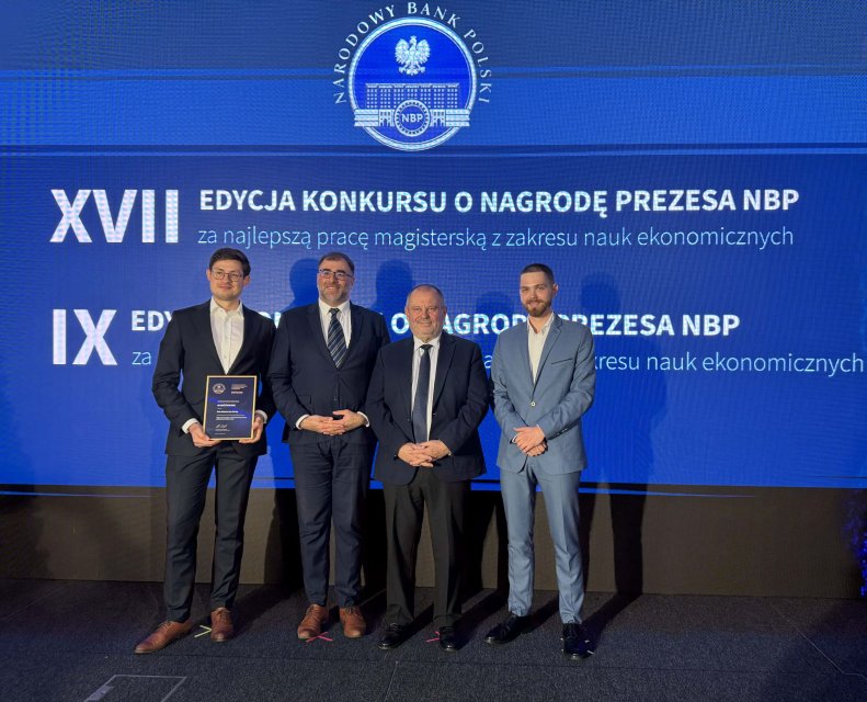 Na zdjęciu przedstawiono formalne wydarzenie, prawdopodobnie ceremonię wręczenia nagrody. Na scenie stoją czterej mężczyźni ubrani w garnitury, na tle dużego ekranu z logo Narodowego Banku Polskiego oraz napisem informującym o edycji konkursu o Nagrodę Prezesa NBP za najlepszą pracę magisterską z zakresu ekonomii. Pierwszy mężczyzna od lewej trzyma oprawiony certyfikat, ma na sobie ciemny garnitur, jasną koszulę i okulary. Obok niego stoi mężczyzna w ciemnym garniturze, z krawatem i okularami. Pośrodku znajduje się kolejny mężczyzna w ciemnym garniturze i krawacie. Po prawej stronie stoi mężczyzna w jasnoniebieskim garniturze i białej koszuli, ogolony, bez zarostu. Tło utrzymane jest w odcieniach niebieskiego, co podkreśla oficjalny charakter wydarzenia. Oświetlenie skierowane jest na uczestników i dyplom, tworząc delikatne cienie, a kompozycja zdjęcia jest prosta, z postaciami umieszczonymi centralnie na tle ekranu.