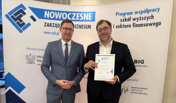 Zdjęcie przedstawia dwóch mężczyzn ubranych w garnitury. Mężczyzna po prawej trzyma dyplom. Oba mężczyźni uśmiechają się. W tle na banerze widać napisy: Nowoczesne Zarządzanie Biznesem.