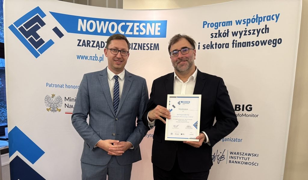 Zdjęcie przedstawia dwóch mężczyzn ubranych w garnitury. Mężczyzna po prawej trzyma dyplom. Oba mężczyźni uśmiechają się. W tle na banerze widać napisy: Nowoczesne Zarządzanie Biznesem.