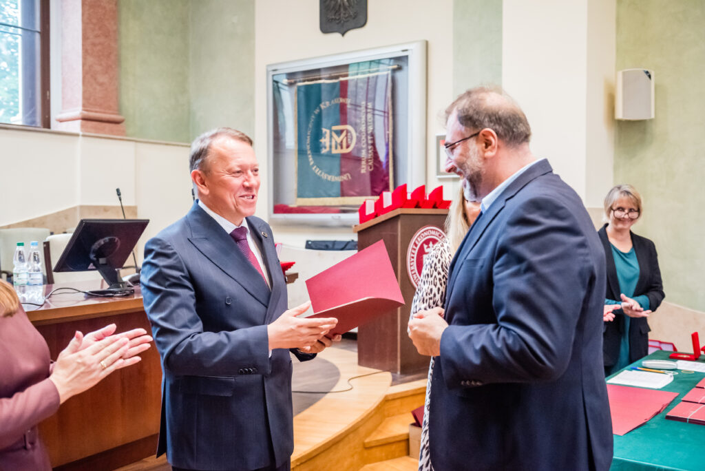 Wręczenie dokumentów podczas uroczystości akademickiej w sali reprezentacyjnej uczelni.