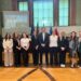 Na zdjęciu znajduje się grupa jedenastu osób ustawionych w jednym rzędzie w przestronnej sali konferencyjnej. W tle widoczne są duże, prostokątne okna oraz ekran projekcyjny, na którym wyświetlona jest prezentacja z napisem „BEYOND THE DECADES: CELEBRATING A JOINT HISTORY, INSPIRING THE FUTURE”. Za grupą stoją flagi, w tym flaga Unii Europejskiej oraz Polski. Po prawej stronie widoczna jest mównica z logo uczelni. Podłoga wykonana jest z jasnego drewna, a całość wnętrza utrzymana jest w eleganckim, oficjalnym stylu.