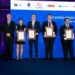 Zdjęcie grupowe laureatów i organizatorów X polskiej edycji Konkursu 'Etyka w finansach'. Pięć osób w centralnej części kadru trzyma oprawione dyplomy, stojąc na tle niebieskiej ścianki z logotypami partnerów konkursu, m.in. Związku Banków Polskich, Banku Pekao, PKO Banku Polskiego, Santander Bank Polska, BNP Paribas i innych. Laureaci ubrani są formalnie – kobieta w eleganckiej czarnej sukience, mężczyźni w garniturach. Po bokach stoją czterej starsi mężczyźni – członkowie jury oraz przedstawiciele instytucji wspierających.