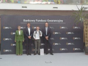 Indywidualne zdjęcie laureata trzymającego dyplom, stojącego na tle ścianki z logo Bankowego Funduszu Gwarancyjnego. Po jego bokach stoją trzej mężczyźni i jedna kobieta w eleganckich strojach. Na podłodze przed laureatem znajdują się dwie białe torby z upominkami.