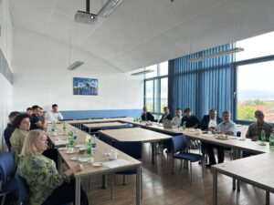 Na pierwszym planie widzimy grupę osób siedzących przy stołach w kształcie litery U. Stoły są ustawione w jasnym pomieszczeniu. Na stołach znajdują się napoje, filiżanki i talerze z przekąskami. Ludzie wyglądają na skupionych. W tle dostrzec można duże okna z niebieskimi zasłonami, przez które widać drzewa i fragment miejskiego krajobrazu, co sugeruje, że pokój znajduje się na wyższych piętrach budynku. Na ścianie widnieje obraz lub plakat przedstawiający fragment natury. Atmosfera jest formalna i profesjonalna. Część osób wygląda na zaangażowanych w rozmowę lub słuchanie prezentacji. Widać również projektor podwieszony na suficie. Pora dnia sugerowana przez oświetlenie i widoczność za oknami to późne rano lub wczesne popołudnie. Pomieszczenie jest dobrze oświetlone naturalnym światłem.
