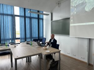 W obrazie przedstawiono nowoczesną salę. Pierwszy plan: - Znajduje się mężczyzna siedzący przy stole, który używa laptopa. - Na stole widoczne są laptopy, kilka butelek z wodą, filiżanki oraz talerzyki z jakimś jedzeniem. - W tle widzimy duże niebieskie żaluzje zasłaniające okna, przez które przechodzi światło dzienne. Na ścianie znajduje się tablica oraz ekran, na którym jest wyświetlany pulpit z projektora. - Jasność obrazu sugeruje, że zdjęcie zostało zrobione w ciągu dnia. Pokój jest przestronny i dobrze oświetlony naturalnym światłem.