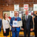 W przedstawionym obrazie możemy zobaczyć grupę pięciu osób stojących w pomieszczeniu o wyglądzie muzealnym. W centralnej części obrazu znajduje się mężczyzna trzymający w rękach dyplom. Wokół niego znajdują się cztery inne osoby - dwie z lewej i dwie z prawej strony. Tło stanowią ceglaste ściany z zawieszonymi dokumentami i fotografiami, co sugeruje, że miejsce to ma charakter historyczny lub edukacyjny. W tle widoczne są symbole takie jak godło Polski, co może wskazywać na oficjalny charakter instytucji. Osoby na zdjęciu są ubrane w eleganckie stroje. Wszyscy patrzą w stronę aparatu, a ich wyraz twarzy jest profesjonalny, co wskazuje na oficjalne spotkanie bądź ceremonię wręczenia nagród. Atmosfera obrazu jest formalna i profesjonalna. Wystrój pomieszczenia oraz obecność dokumentów wskazują na instytucjonalność miejsca. Czas dnia nie jest ściśle określony, ale jasne oświetlenie sugeruje, że zdjęcie mogło być wykonane w ciągu dnia.
