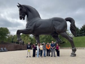 Na zdjęciu znajduje się grupa ośmiu osób ustawionych w szeregu na tle monumentalnej, ciemnej rzeźby konia w dynamicznej pozie. Postument z figurą stoi na utwardzonym, jasnym placu, a w tle widoczne są drzewa i zielone wzgórze. Osoby mają na sobie codzienne stroje, w rękach trzymają torby oraz dokumenty. Niebo jest pochmurne, co podkreśla monumentalny charakter rzeźby.