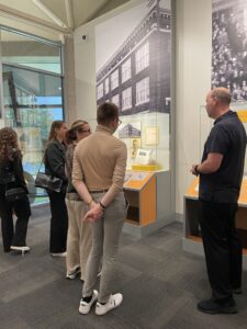 Na zdjęciu znajduje się kilka osób zwiedzających budynek muzeum. Grupa stoi przed witrynami z eksponatami oraz planszami informacyjnymi, poświęconymi architekturze oraz wydarzeniom z przeszłości. Osoby w centrum kadru są zwrócone twarzami do gablot, rozmawiając lub czytając opisy. W tle widoczne są duże czarno-białe zdjęcia budynków i tłumu ludzi, a po lewej stronie znajduje się przeszklona ściana, przez którą wpada naturalne światło. Pomieszczenie urządzone jest w nowoczesnym stylu, z szarymi wykładzinami i przeszklonymi elementami.