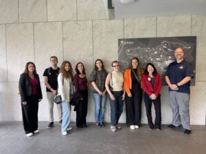 Na zdjęciu znajduje się grupa dziewięciu osób stojących w rzędzie na tle jasnej ściany oraz dużej mapy ekspozycji “Frederik Meijer Gardens & Sculpture Park”. Osoby ubrane są w casualowe stroje – widoczne są marynarki, bluzy, swetry oraz codzienne obuwie. Większość z nich ma przy sobie torby lub plecaki. Pomieszczenie jest przestronne, z szarą podłogą i minimalistycznym wystrojem. Cała grupa pozuje razem na pamiątkowym zdjęciu.