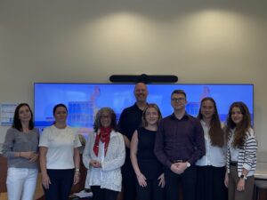 Na zdjęciu znajduje się grupa ośmiu osób stojących w pomieszczeniu biurowym, biorących udział w spotkaniu z DIANA LAWSON – Dean, Seidman College of Business. W tle widoczny jest duży ekran z niebieskim tłem przedstawiającym nowoczesny budynek. Osoby ustawione są w dwóch rzędach — większość stoi z przodu, a jedna osoba znajduje się z tyłu, pośrodku grupy. Pomieszczenie jest jasne i uporządkowane, z beżowymi ścianami i prostymi meblami biurowymi.