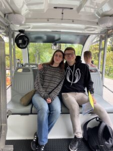 Na zdjęciu znajdują się dwie osoby siedzące obok siebie w pojeździe typu meleks. Osoba po lewej stronie ma na sobie sweter w paski i dżinsy, a osoba po prawej czarną bluzę z białym wzorem oraz jasne spodnie. Obie osoby uśmiechają się i obejmują ramionami. W tle, przez okna pojazdu, widoczna jest zielona roślinność. Przed siedzącymi osobami leży czarny plecak i żółta kartka. W pojeździe panuje jasne, naturalne światło.