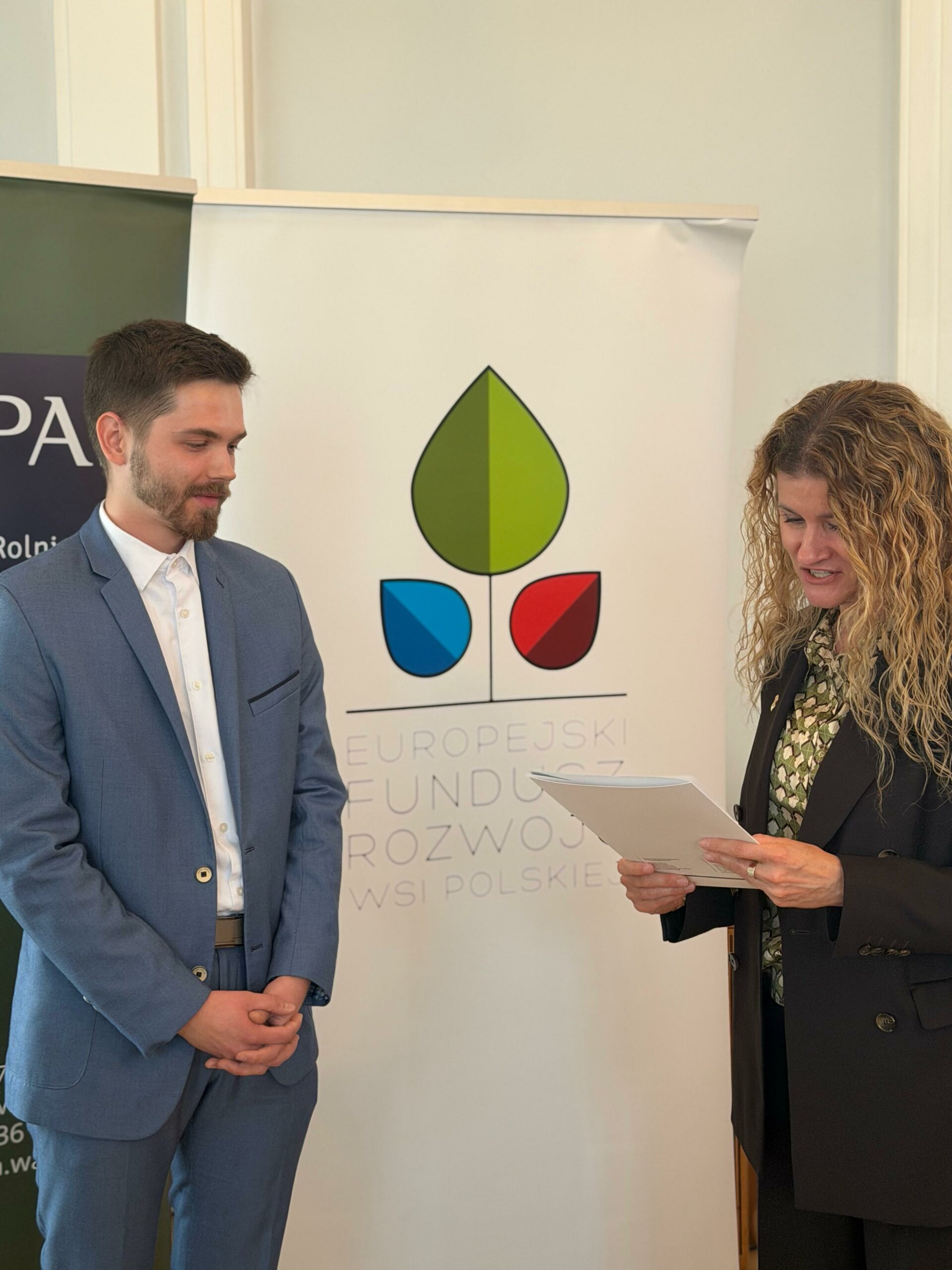 Na zdjęciu widać mężczyznę i kobietę stojących w pomieszczeniu podczas formalnego wydarzenia lub konferencji. Mężczyzna po lewej ma jasnoniebieski garnitur i białą koszulę, trzyma dłonie przed sobą i z lekkim uśmiechem patrzy na kobietę. Kobieta po prawej ma długie, kręcone blond włosy, ciemną marynarkę i trzyma w rękach plik dokumentów, jakby prowadziła prezentację lub rozmowę. W tle widoczny jest baner z logo i napisem „Europejski Fundusz Rozwoju Wsi Polskiej”, co sugeruje, że wydarzenie dotyczy rozwoju obszarów wiejskich. Atmosfera jest profesjonalna i oficjalna, oświetlenie jasne i równomierne.