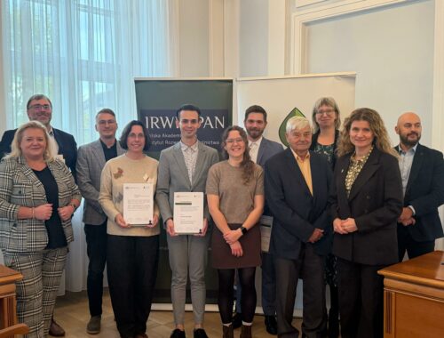 Na zdjęciu widać grupę dziesięciu osób pozujących do wspólnej fotografii w pomieszczeniu, prawdopodobnie podczas oficjalnego wydarzenia lub ceremonii wręczenia nagród. Uczestnicy — kobiety i mężczyźni w eleganckich strojach — stoją w dwóch rzędach i patrzą w obiektyw, uśmiechając się. W centrum znajdują się dwie osoby trzymające certyfikaty, co sugeruje wyróżnienie lub zakończenie projektu. W tle widoczny jest baner z napisem „IRW PAN” i „Polska Akademia Nauk”, wskazujący na związek wydarzenia z instytucją naukową. Atmosfera zdjęcia jest uroczysta, ale jednocześnie przyjazna i pełna dumy.