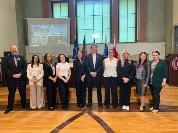 Na zdjęciu znajduje się grupa jedenastu osób ustawionych w jednym rzędzie w przestronnej sali konferencyjnej. W tle widoczne są duże, prostokątne okna oraz ekran projekcyjny, na którym wyświetlona jest prezentacja z napisem „BEYOND THE DECADES: CELEBRATING A JOINT HISTORY, INSPIRING THE FUTURE”. Za grupą stoją flagi, w tym flaga Unii Europejskiej oraz Polski. Po prawej stronie widoczna jest mównica z logo uczelni. Podłoga wykonana jest z jasnego drewna, a całość wnętrza utrzymana jest w eleganckim, oficjalnym stylu.