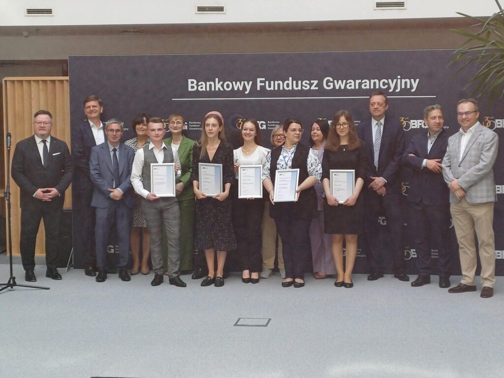 Grupowe zdjęcie uczestników i organizatorów wydarzenia z okazji wręczenia nagród lub certyfikatów przez Bankowy Fundusz Gwarancyjny. W pierwszym rzędzie stoi sześć osób trzymających dyplomy, za nimi grupa przedstawicieli w formalnych strojach. W tle ścianka z logo Bankowego Funduszu Gwarancyjnego.