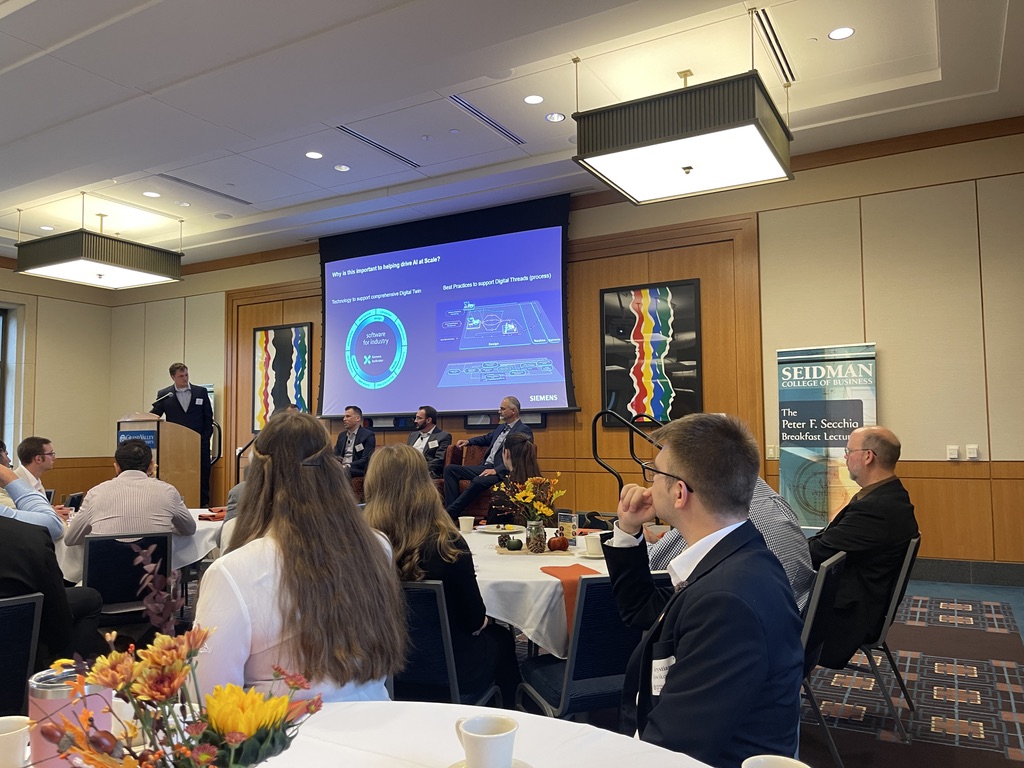 Na zdjęciu uchwycona jest scena z konferencji lub wykładu w nowoczesnej, jasnej sali. Na pierwszym planie widać kilka okrągłych stołów, przy których siedzą uczestnicy wydarzenia. Przed salą, na podwyższeniu, znajduje się panelistów – cztery osoby siedzące oraz jedna stojąca przy mównicy, prowadząca prezentację widoczną na dużym ekranie. Temat prezentacji dotyczy nowoczesnej technologii, co sugeruje wyświetlana plansza z logo Siemens. W tle widoczne są banery Seidman College of Business, kwiatowe dekoracje na stołach oraz obrazy na ścianie. Panuje formalna atmosfera wydarzenia.