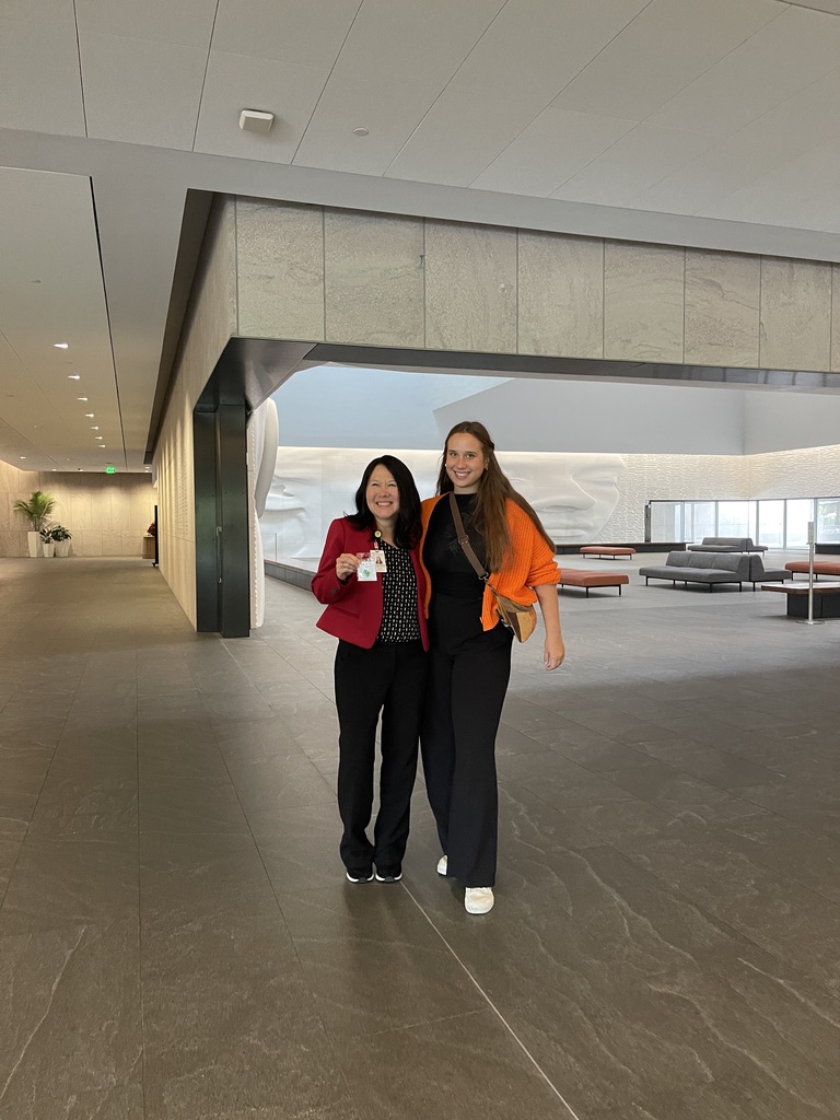 Na zdjęciu znajdują się dwie kobiety stojące razem w nowoczesnym i przestronnym wnętrzu budynku. Kobieta po lewej ubrana jest w czerwony żakiet i czarne spodnie, trzyma w ręku identyfikator. Kobieta po prawej ma na sobie czarny strój z pomarańczowym swetrem przewiązanym na ramionach. W tle widoczna jest minimalistyczna przestrzeń z jasnymi ścianami, designerskimi ławkami i dużą, białą rzeźbą na ścianie. Wnętrze jest dobrze oświetlone, a po lewej stronie kadru ciągnie się korytarz.