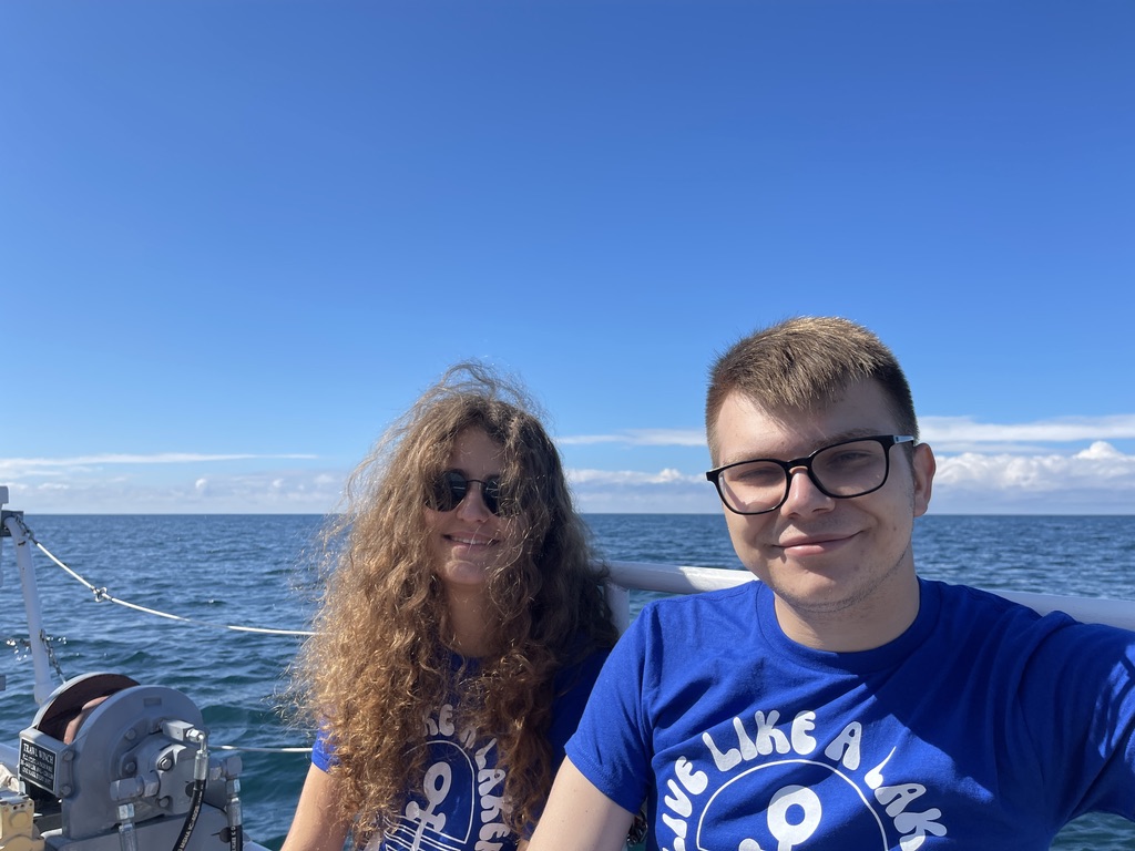 Na zdjęciu znajdują się dwie osoby siedzące na pokładzie łodzi na tle rozległego jeziora Michigan. Obydwie mają na sobie niebieskie koszulki z białym napisem i symbolem kotwicy. Na dalszym planie widoczna jest linia horyzontu oraz niebo z pojedynczymi chmurami, a w dolnym lewym rogu zdjęcia widać fragment sprzętu pokładowego łodzi. Scena odbywa się w słoneczny dzień, a atmosfera zdjęcia jest letnia i spokojna.