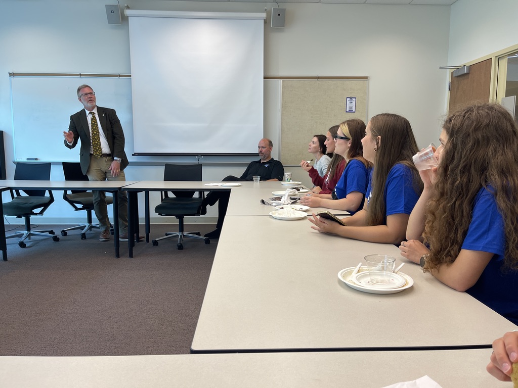 Na zdjęciu znajduje się grupa osób uczestniczących w spotkaniu lub wykładzie w sali konferencyjnej. Po prawej stronie stołu siedzi kilka młodych osób w niebieskich koszulkach, jedząc i pijąc napoje. Po lewej stronie kadru, przed tablicą i ekranem projekcyjnym, stoi mężczyzna w garniturze - MARK SCHAUB – Dean Brooks College of Interdisciplinary Studies, który prowadzi prezentację lub dyskusję. W tle widać krzesła konferencyjne oraz minimalistyczny wystrój pomieszczenia. Atmosfera spotkania jest formalna, a stoły ustawione są w kształcie litery U.