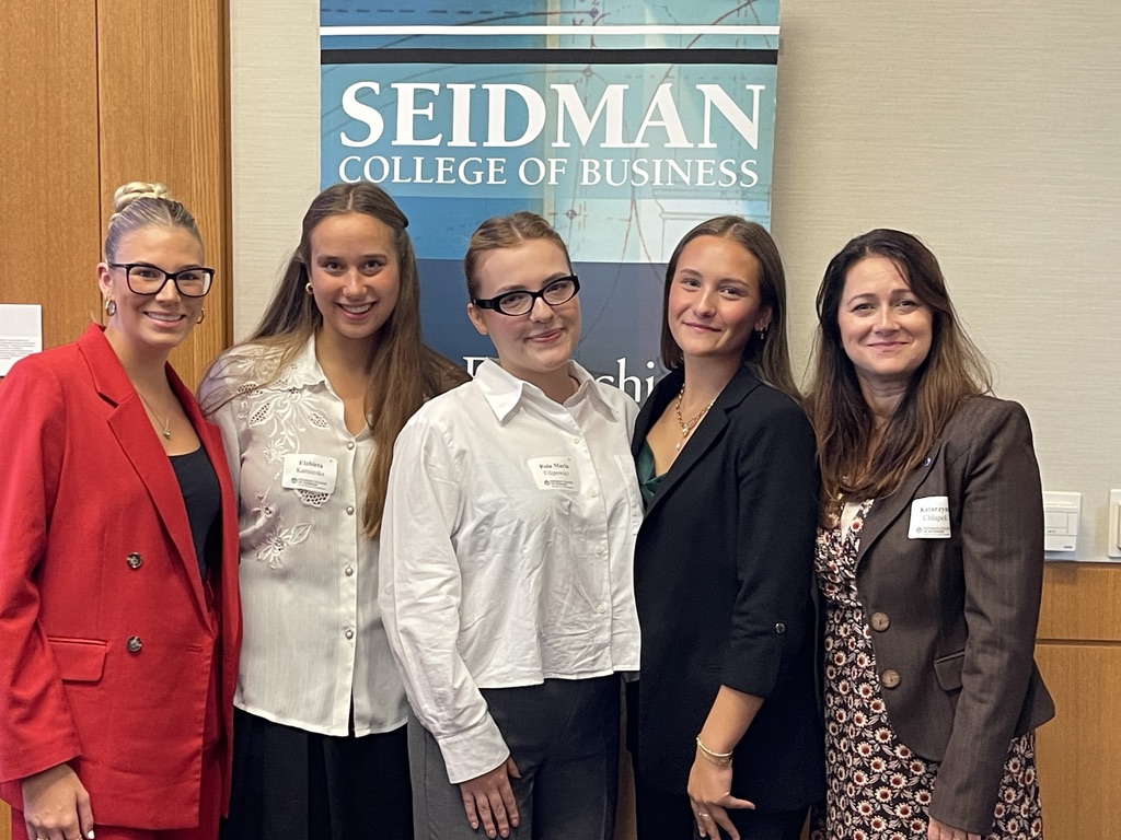 Na zdjęciu znajduje się grupa pięciu kobiet stojących razem na tle baneru z napisem “Seidman College of Business”. Wszystkie osoby ubrane są elegancko: widoczne są marynarki, żakiety oraz sukienka w kwiaty. Część osób ma przypięte identyfikatory, co wskazuje na udział w konferencji. Tło stanowią jasne ściany oraz fragment drewnianej boazerii, nadające przestrzeni profesjonalny charakter.