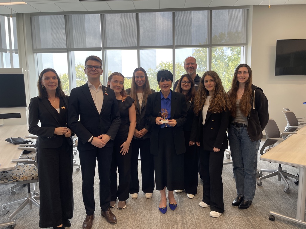 Na zdjęciu znajduje się grupa dziesięciu osób stojących w nowoczesnej sali konferencyjnej. W centrum kadru stoi kobieta - Prezydent GVSU - PHILOMENA V. MANTELLA trzymająca w dłoniach statuetkę wręczoną przez delegację UEK. Za grupą widoczne są duże okna, przez które wpada naturalne światło, oraz drzewa na zewnątrz budynku. Po bokach znajdują się stoły i krzesła biurowe, a na ścianie po prawej stronie wisi płaski ekran. Pomieszczenie jest jasne, przestronne i urządzone w nowoczesnym stylu.