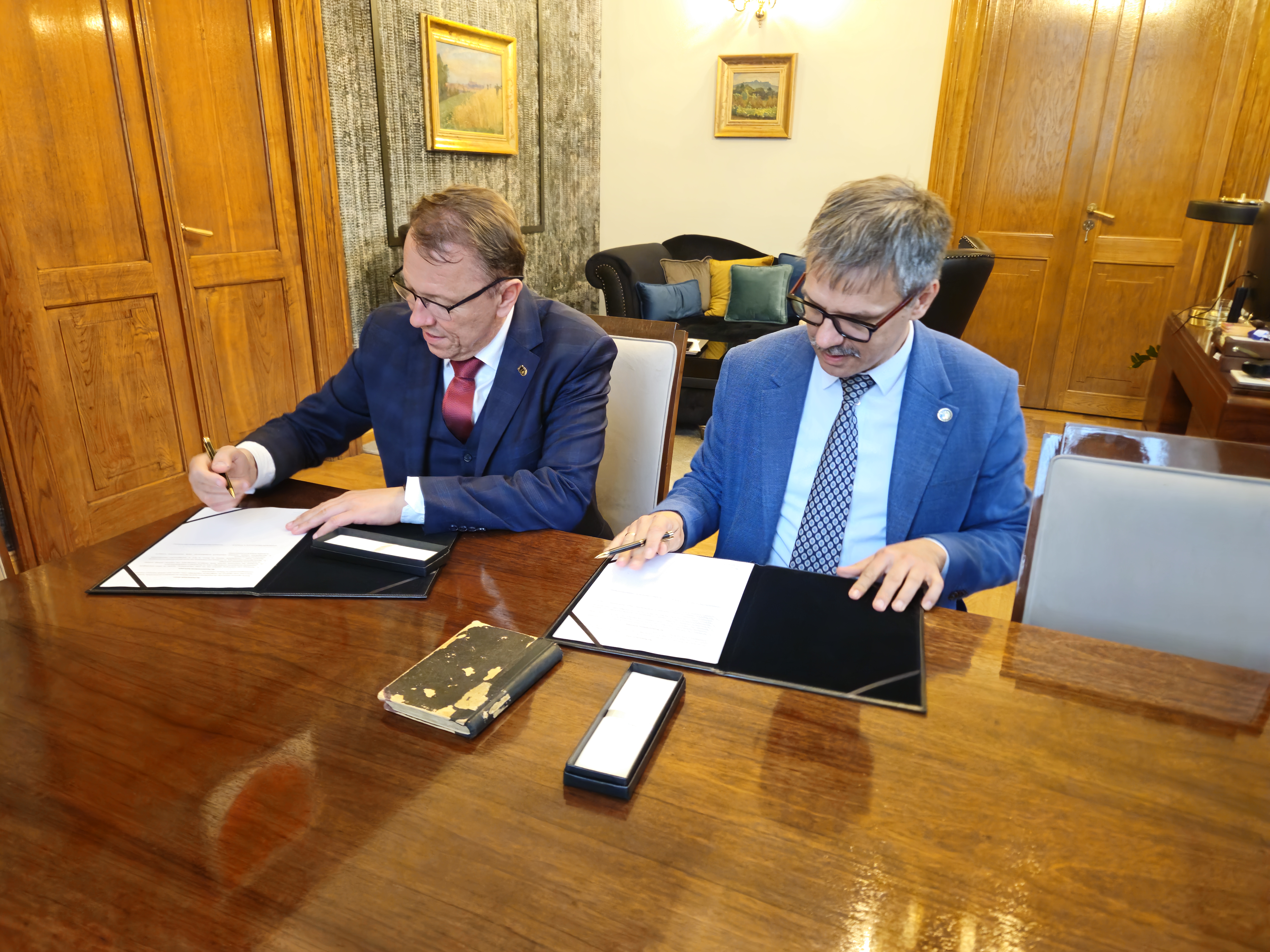 Na pierwszym planie zdjęcia znajdują się dwie osoby siedzące przy dużym, drewnianym stole. Obaj mężczyźni są w garniturach i piszą na dokumentach. Ich wyraz twarzy jest skupiony, co świadczy o powadze sytuacji. W tle pomieszczenia można zobaczyć drewniane drzwi, obrazy na ścianach oraz kanapę z kolorowymi poduszkami. Widać także dekoracyjne światło na suficie, co sugeruje, że jest to elegancki pokój. Atmosfera zdjęcia jest formalna i profesjonalna, z nutą oficjalności i koncentracji.