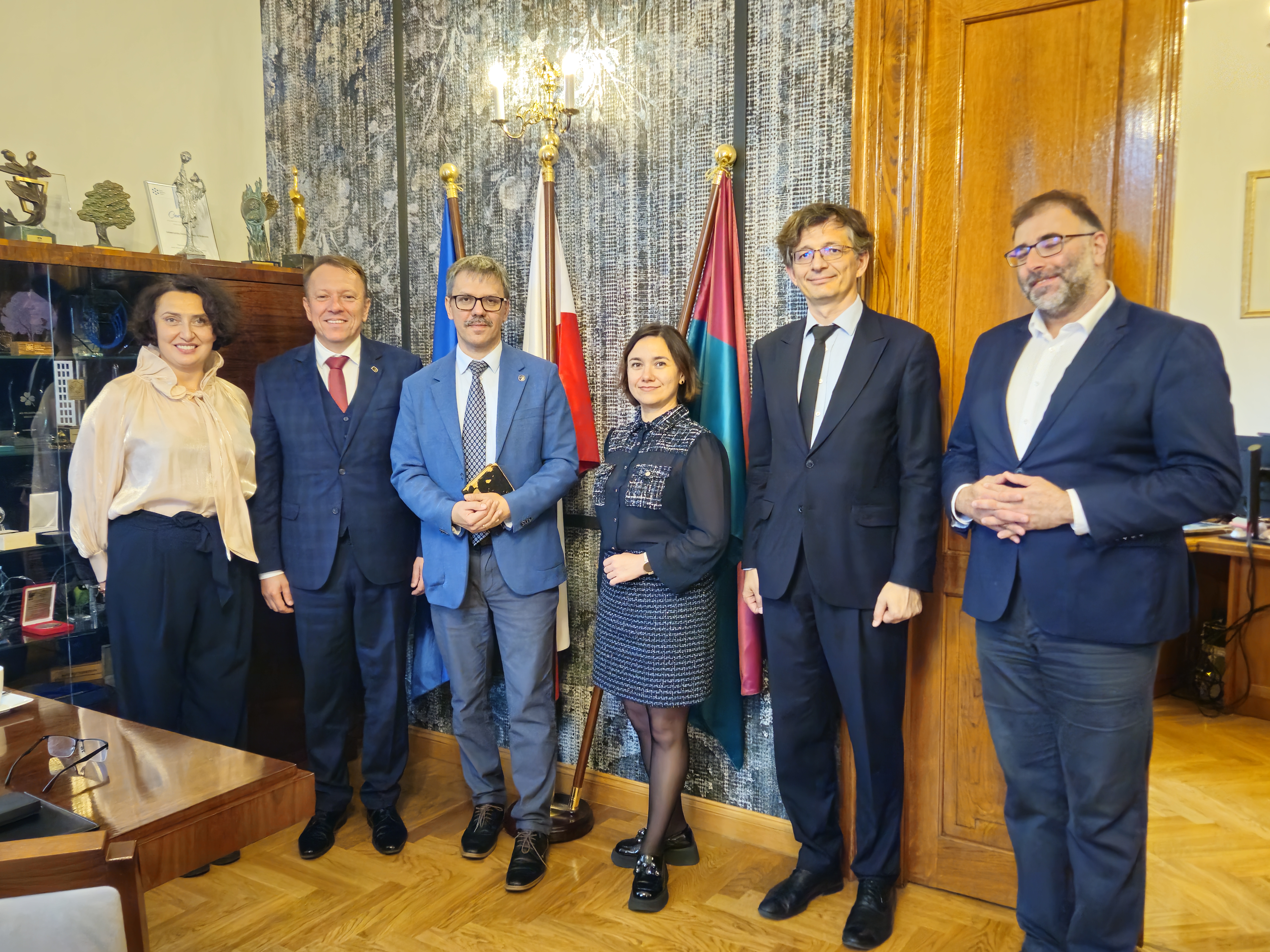 Na zdjęciu widoczne jest sześć osób pozujących do zdjęcia. Wszystkie są elegancko ubrane. W tle widać drewniane meble i zasłony. Na półce znajdują się różne dekoracje i nagrody, a za osobami stoją flagi. Jest także lampa na ścianie, co dodaje elegancji i formalności. Osoby stoją blisko siebie, co wskazuje na przyjazną. Atmosfera wydaje się być formalna, ale przyjazna. Osoby wydają się być w dobrych nastrojach, co można wnioskować z ich postawy i wyrazów twarzy. Wystrój sugeruje eleganckie biuro, co jest podkreślone przez drewniane elementy wystroju oraz obecność flag. Nie ma wyraźnych wskazówek dotyczących pory dnia ani roku, aczkolwiek sztuczne oświetlenie może sugerować wnętrze niezależne od pory dnia.