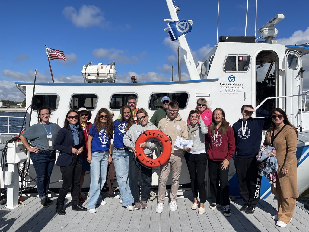 Na zdjęciu znajduje się grupa trzynastu osób ustawionych na pomoście przed dużą, białą łodzią instytutu Grand Valley State University. Uczestnicy są ubrani w swobodne stroje, część z nich ma niebieskie lub bordowe bluzy z logotypem, a kilka osób trzyma certyfikaty lub pomarańczowe koło ratunkowe z napisem “GVSU”. W tle widać flagę USA, sprzęt ratunkowy oraz wyraźnie niebieskie niebo z pojedynczymi chmurami. Scena jest dynamiczna, uczestnicy uśmiechają się, a atmosfera jest pogodna i oficjalna.