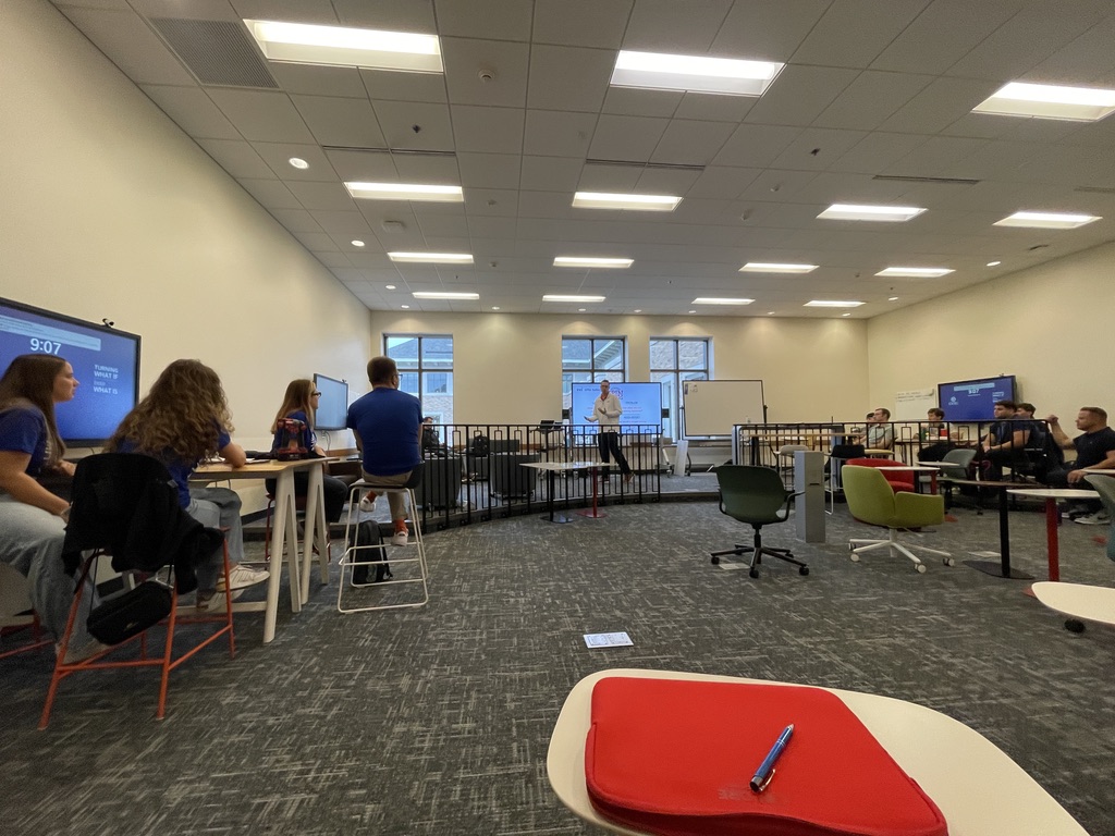 Na zdjęciu znajduje się grupa uczestników siedzących w nowoczesnej, przestronnej sali wykładowej. W centrum kadru, za niską barierką, znajduje się osoba prowadząca zajęcia, stojąca na tle tablicy i dużych okien, przez które wpada naturalne światło. Po obu stronach pomieszczenia znajdują się ekrany wyświetlające prezentacje oraz białe tablice z notatkami. Pomieszczenie wyposażone jest w różnorodne kolorowe krzesła i stoliki; na pierwszym planie widać biały stolik z czerwonym etui oraz długopisem. Atmosfera jest robocza, a ustawienie sali sprzyja dyskusjom grupowym.