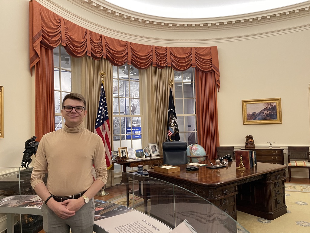 Na zdjęciu znajduje się mężczyzna stojący w zrekonstruowanym wnętrzu Gabinetu Owalnego, charakterystycznego dla Białego Domu. Pomieszczenie wyróżnia się eleganckimi zasłonami w ceglastym kolorze, ciemnym drewnianym biurkiem prezydenckim z licznymi pamiątkami, książkami i globusem. W tle widoczne są dwie flagi – Stanów Zjednoczonych oraz prezydencka – oraz duże okna ze szprosami. Na ścianach zawieszony jest obraz oraz fotografie na stole przy oknie. Całość wnętrza podkreśla oficjalny, prestiżowy charakter miejsca.