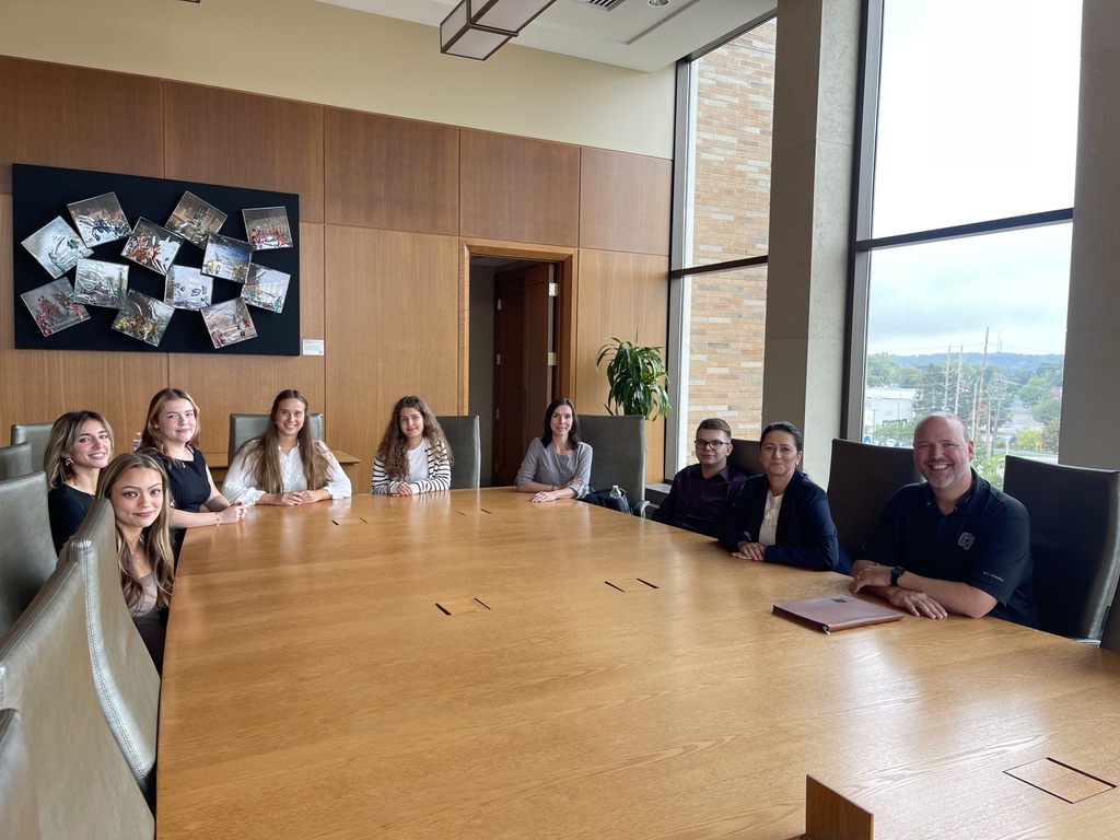 Na zdjęciu znajduje się grupa dziewięciu osób siedzących wokół dużego, drewnianego stołu konferencyjnego w przestronnej sali spotkań. Pokój ma duże okna, przez które wpada naturalne światło i widać panoramę miasta oraz fragment nowoczesnego budynku. Ściany sali ozdobione są jasnym drewnem oraz zdjęciami w czarnych ramkach, a w rogu stoi zielona roślina doniczkowa. Osoby przy stole są elegancko ubrane, co podkreśla formalny charakter spotkania.