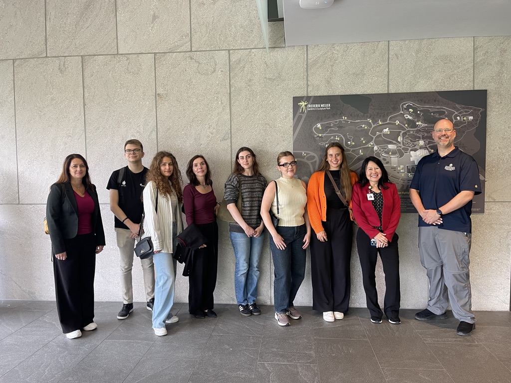 Na zdjęciu znajduje się grupa dziewięciu osób stojących w rzędzie na tle jasnej ściany oraz dużej mapy ekspozycji “Frederik Meijer Gardens & Sculpture Park”. Osoby ubrane są w casualowe stroje – widoczne są marynarki, bluzy, swetry oraz codzienne obuwie. Większość z nich ma przy sobie torby lub plecaki. Pomieszczenie jest przestronne, z szarą podłogą i minimalistycznym wystrojem. Cała grupa pozuje razem na pamiątkowym zdjęciu.
