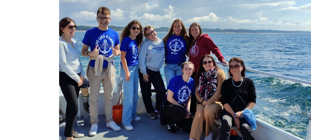 Na zdjęciu znajduje się grupa dziewięciu osób na pokładzie łodzi podczas rejsu. Pięć osób stoi z tyłu, w tle widać rozległą taflę wody i zielony brzeg po prawej stronie. Cztery osoby siedzą z przodu na ławce lub pokładzie, dwie z nich mają na sobie chustki. Kilka osób ubranych jest w niebieskie koszulki z nadrukiem „Live like a lake” i symbolem kotwicy, pozostałe w casualowe stroje. Scena ma charakter pamiątkowy, tło stanowi otwarte jezioro lub morze, widać lekkie fale i zachmurzone, ale jasne niebo.