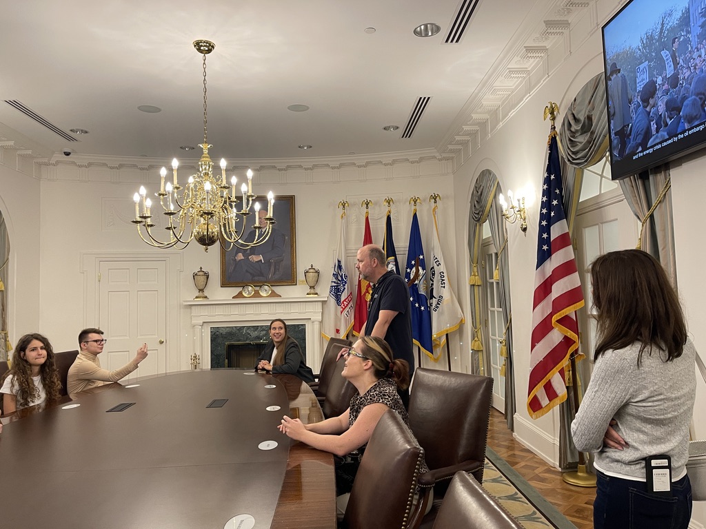 Na zdjęciu widoczna jest grupa osób w eleganckiej sali konferencyjnej, przypominającej wnętrze Białego Domu. Uczestnicy siedzą przy owalnym, ciemnym stole, a po drugiej stronie sali stoi mężczyzna prowadzący spotkanie. W tle znajdują się liczne flagi amerykańskie oraz wojskowe, okazały żyrandol, kominek i portret. Po prawej stronie sali stoi kobieta z założonymi rękami, a na ścianie wisi telewizor wyświetlający obraz demonstracji. Całość przestrzeni podkreśla oficjalny, prestiżowy charakter wnętrza.