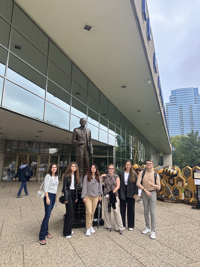 Na zdjęciu znajduje się grupa siedmiu osób pozujących przy pomniku postaci stojącej przed nowoczesnym, przeszklonym budynkiem. Osoby są ubrane w casualowe i półformalnie stroje, takie jak jeansy, marynarki oraz swetry. W tle widoczna jest fasada budynku z dużymi oknami odbijającymi otoczenie, wejście główne oraz element dekoracyjny przypominający strukturę plastra miodu. Fragment miasta z wysokimi budynkami można dostrzec po prawej stronie zdjęcia. Całość sugeruje wizytę studyjną lub wycieczkę w przestrzeni miejskiej.