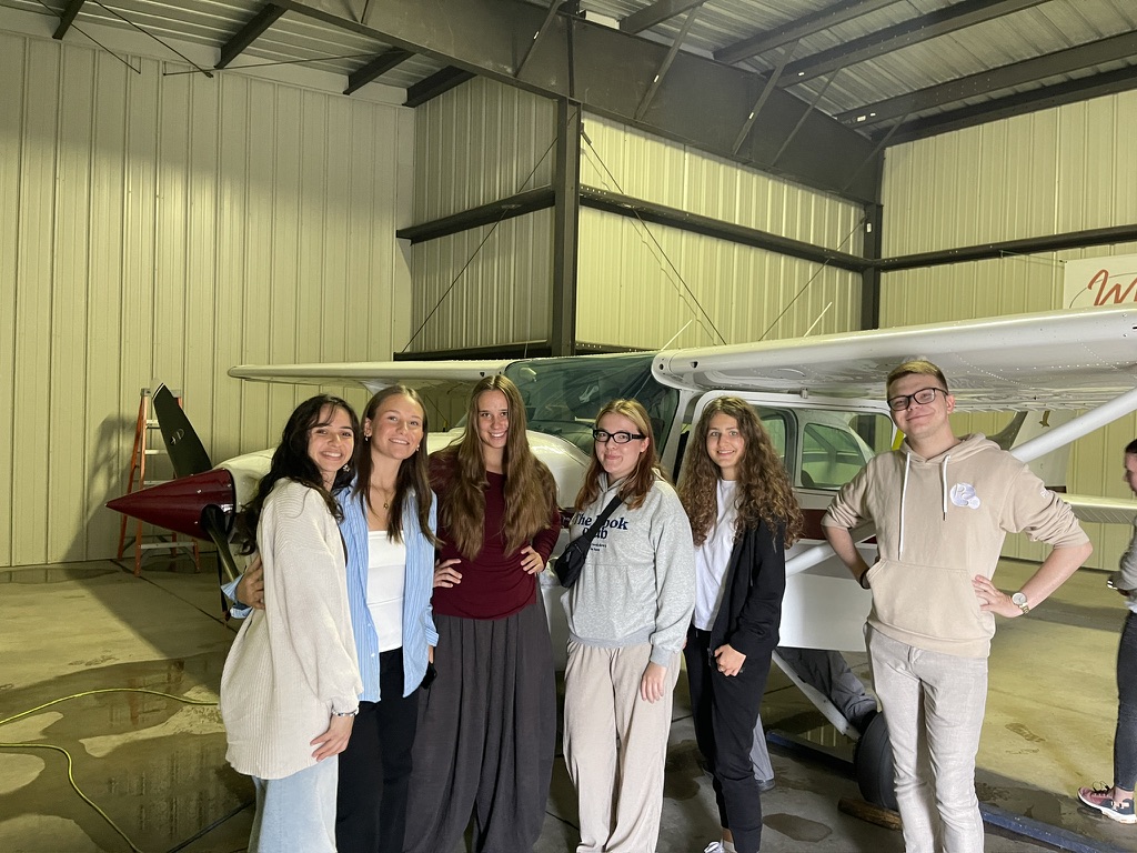 Na zdjęciu znajduje się grupa sześciu osób w hangarze lotniczym. Wszyscy stoją blisko siebie przed niewielkim samolotem, który zajmuje centralną część kadru. Pomieszczenie jest przestronne, z wysokim metalowym sufitem i ścianami w jasnym kolorze. Po lewej stronie widoczna jest drabina oraz fragment skrzydła samolotu o bordowo-białych barwach. Podłoga jest betonowa, a światło wydaje się być sztuczne, rozjaśniające wnętrze hangaru.