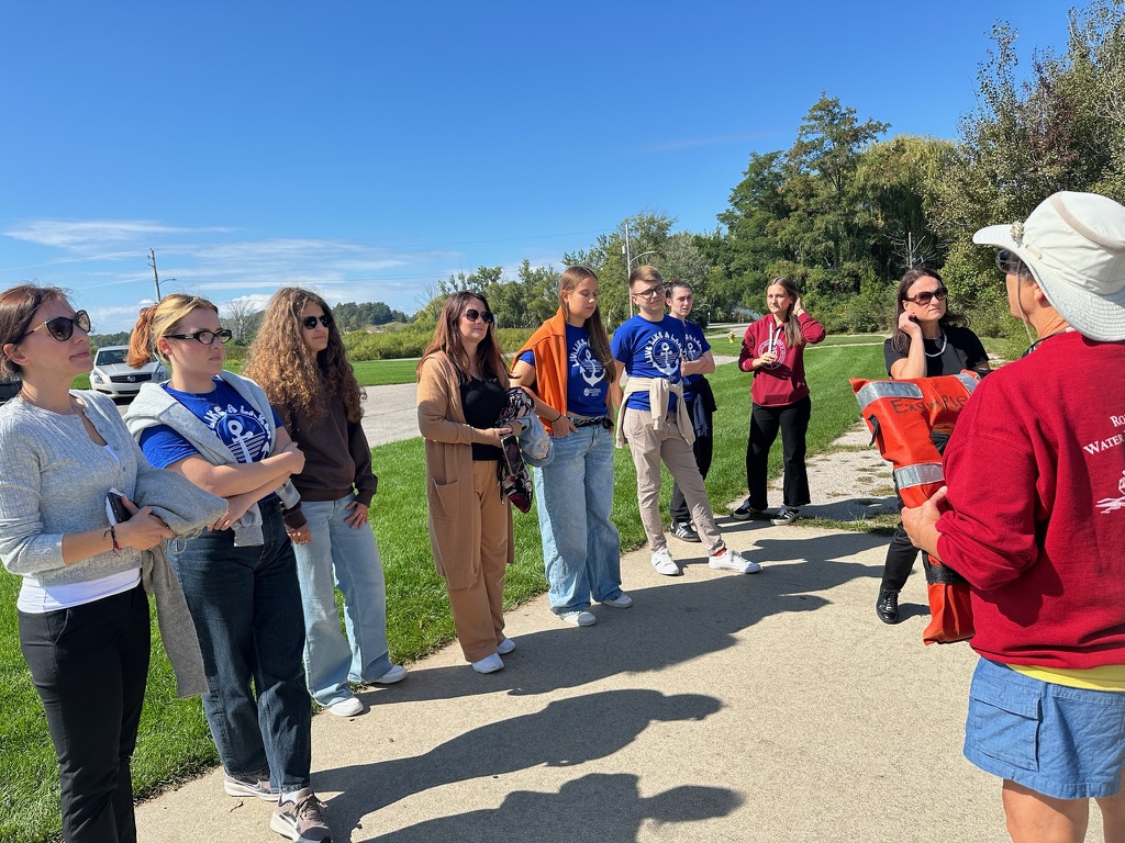Na zdjęciu znajduje się grupa jedenastu osób na zewnątrz, podczas słonecznego dnia. Osoby stoją w półokręgu na chodniku, zwrócone są w kierunku kobiety ubranej w czerwoną bluzę i kapelusz, która trzyma pomarańczową kamizelkę ratunkową. Kilka osób ma na sobie niebieskie koszulki z białym nadrukiem, pozostali noszą swobodne stroje w jasnych i ciemnych kolorach. W tle widoczna jest trawa, drzewa oraz fragment asfaltowej drogi i samochód, a niebo jest bezchmurne i intensywnie niebieskie.