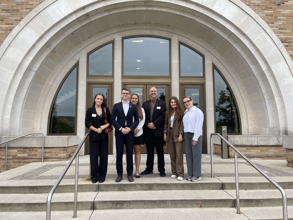 Na zdjęciu znajduje się grupa jedenastu osób na zewnątrz, podczas słonecznego dnia. Osoby stoją w półokręgu na chodniku, zwrócone są w kierunku kobiety ubranej w czerwoną bluzę i kapelusz, która trzyma pomarańczową kamizelkę ratunkową. Kilka osób ma na sobie niebieskie koszulki z białym nadrukiem, pozostali noszą swobodne stroje w jasnych i ciemnych kolorach. W tle widoczna jest trawa, drzewa oraz fragment asfaltowej drogi i samochód, a niebo jest bezchmurne i intensywnie niebieskie.