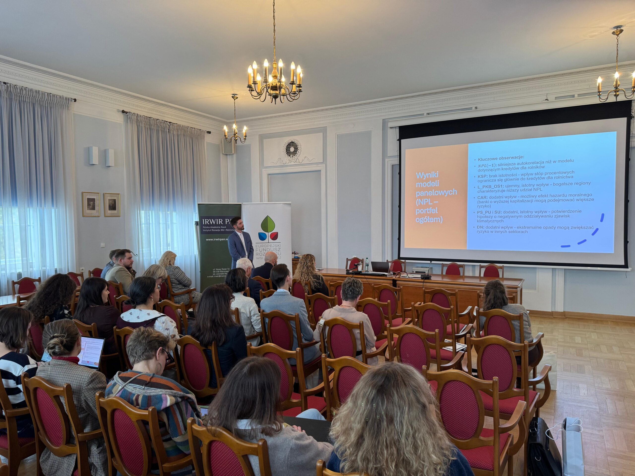 Zdjęcie przedstawia wnętrze sali konferencyjnej o jasnych ścianach i dużych oknach z zasłonami, oświetlone naturalnym światłem. W pierwszym planie widać uczestników siedzących na krzesłach z czerwonymi siedziskami, uważnie słuchających prezentacji — niektórzy robią notatki lub rozmawiają. W tle znajduje się ekran z prezentacją zatytułowaną „Wyniki modeli panelowych (NPL – portfel ogółem)” oraz baner z logo i nazwą „IRWiR PAN”, co wskazuje, że wydarzenie ma charakter naukowy lub biznesowy. Atmosfera jest profesjonalna i skupiona, typowa dla konferencji lub seminarium.